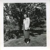 A Japanese American woman, Minnie Nakano, looks on at the camera. She is standing on a lawn in front of a tree.